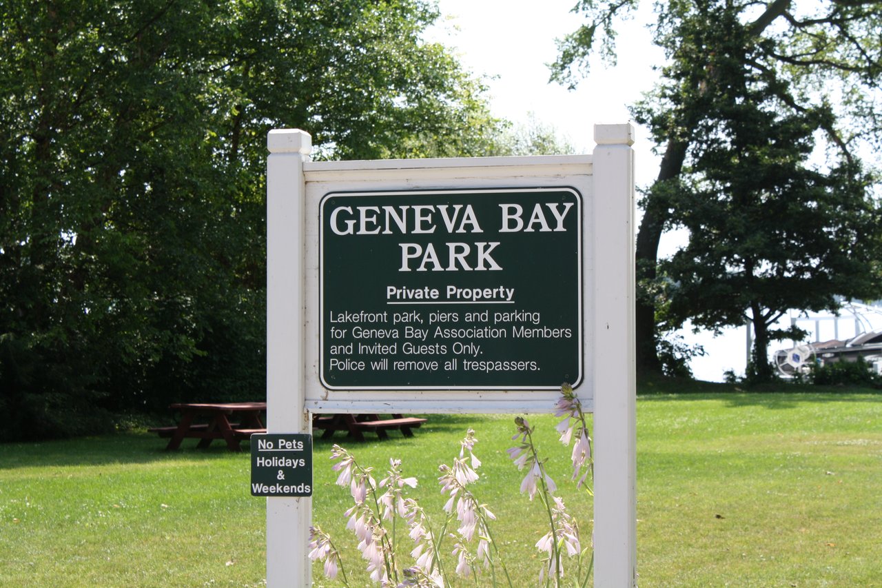 Geneva Bay Park sign beside a grassy lawn, picnic tables, and flowering plants.