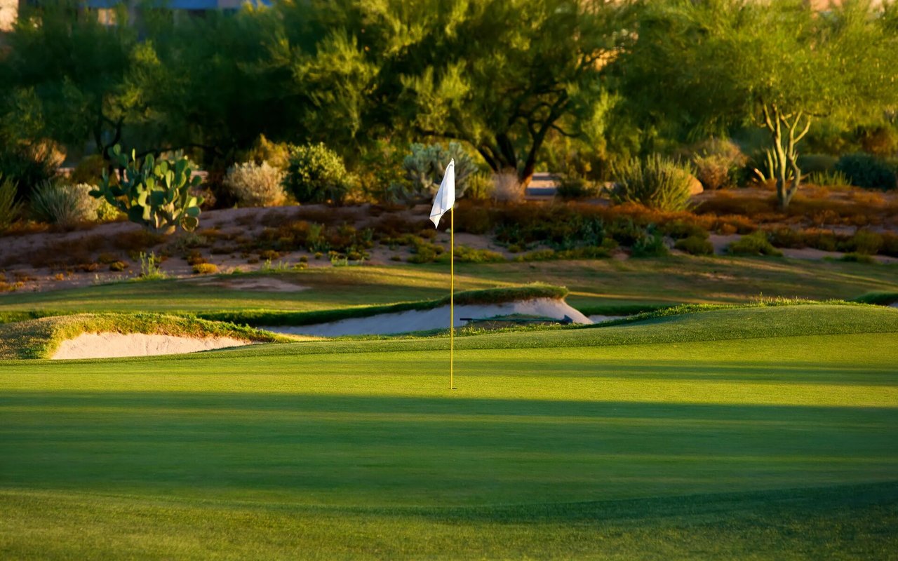 Golf green with a flagstick, sand bunkers, and desert landscaping at golden hour.
