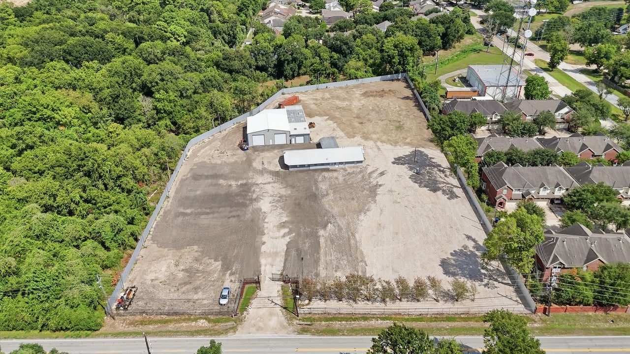 Fenced cleared property with two metal buildings, beside woodland and a residential neighborhood.
