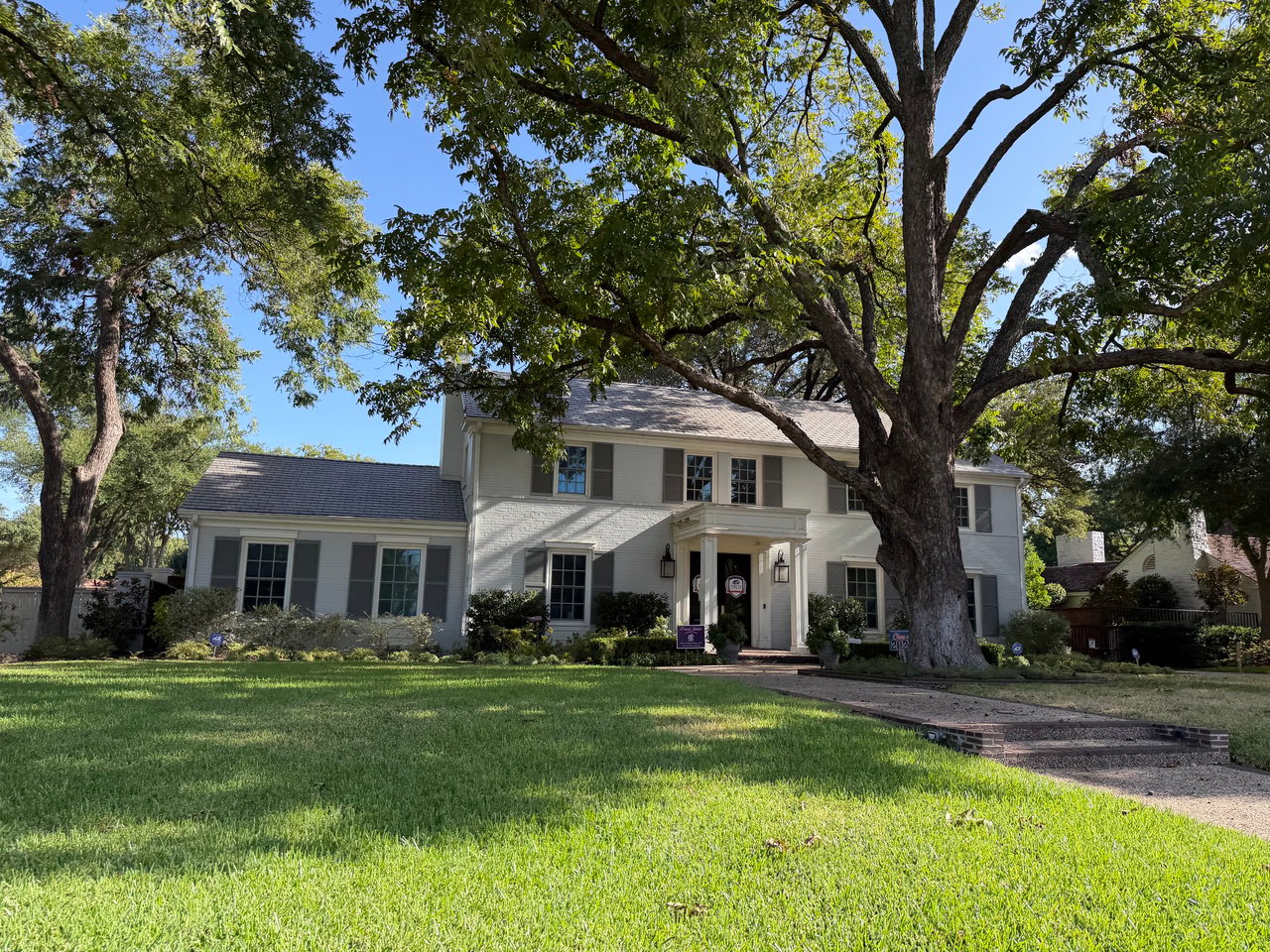 Two-story white house with gray shutters and a front lawn shaded by mature trees.