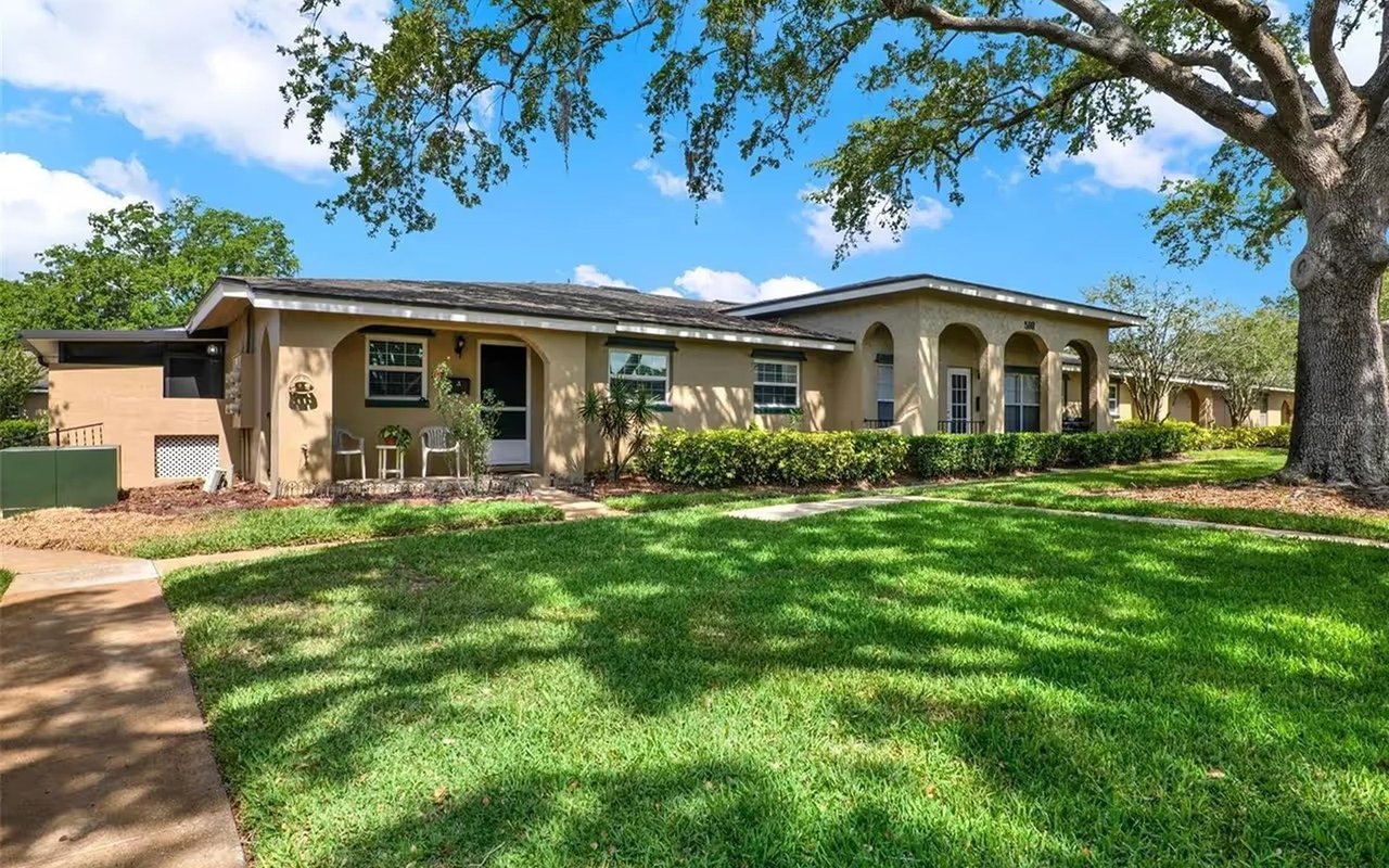 Single-story beige home with an arched entry, broad lawn, and large shade tree.