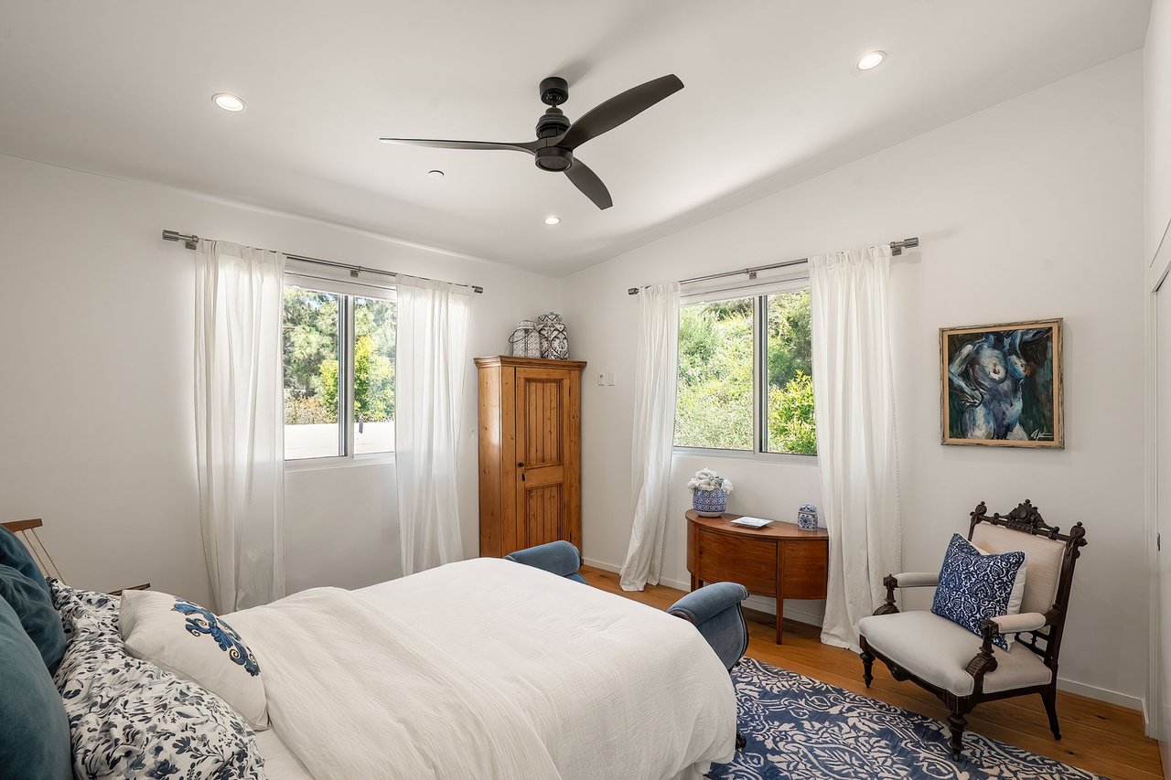 Bedroom with a white bed, blue patterned rug, dark ceiling fan, and windows overlooking greenery.
