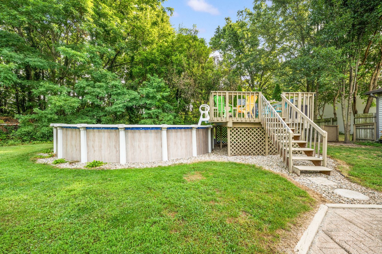 Above-ground pool beside a raised wooden deck with stairs and chairs in a tree-lined backyard.
