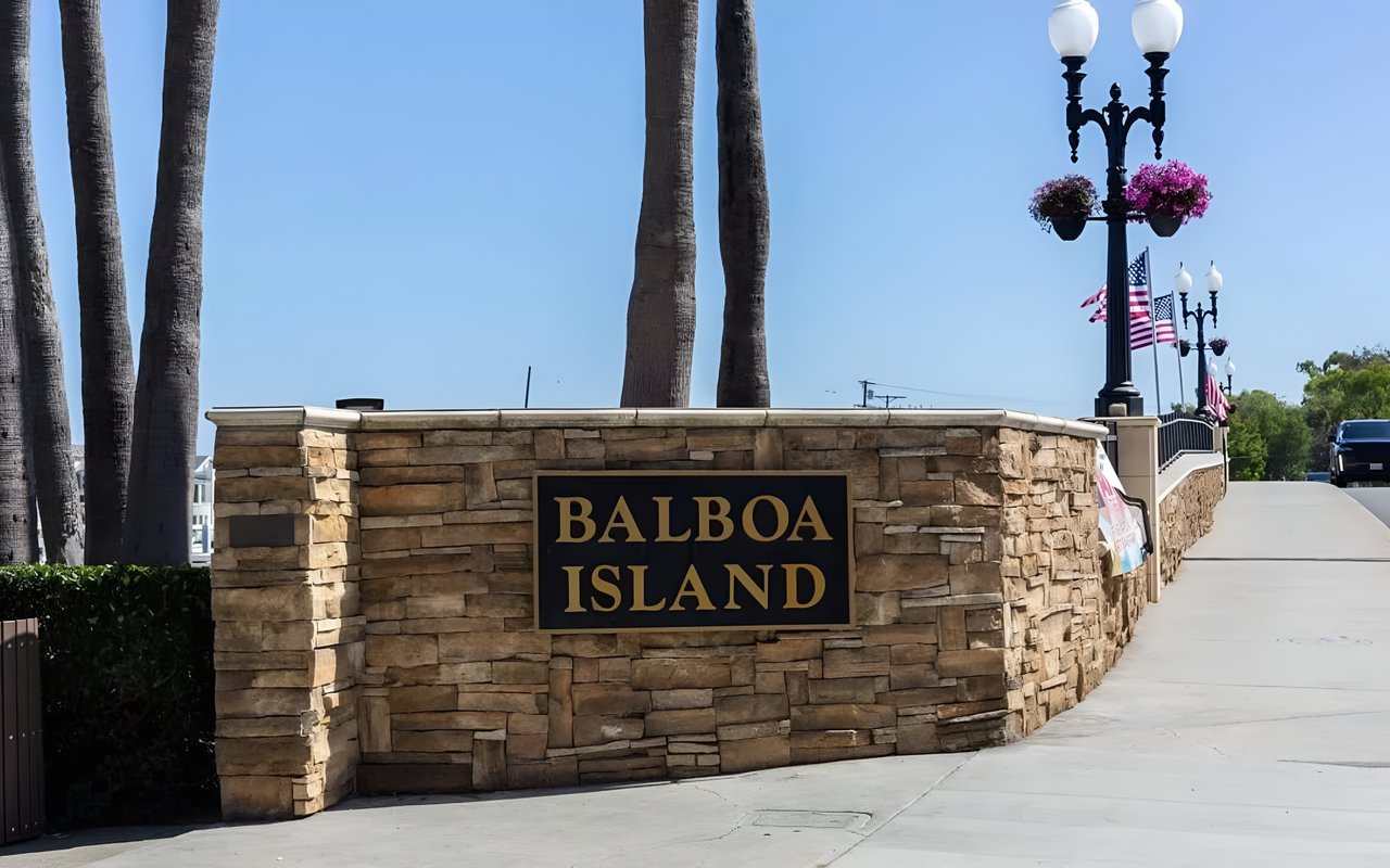  A large stone sign reading "BALBOA ISLAND" stands at the entrance to a bridge with lampposts and palm trees.