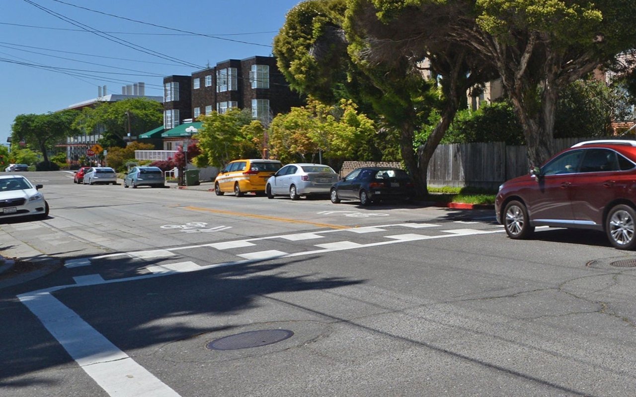 Street intersection with a broad striped crosswalk, parked cars, leafy trees, and low-rise buildings.