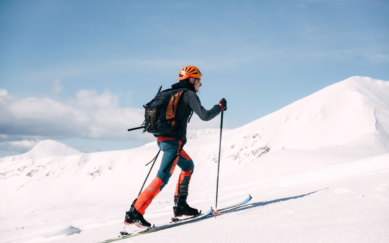 Skier climbs a snowy mountain slope with poles, wearing an orange helmet and bright orange-and-blue gear.