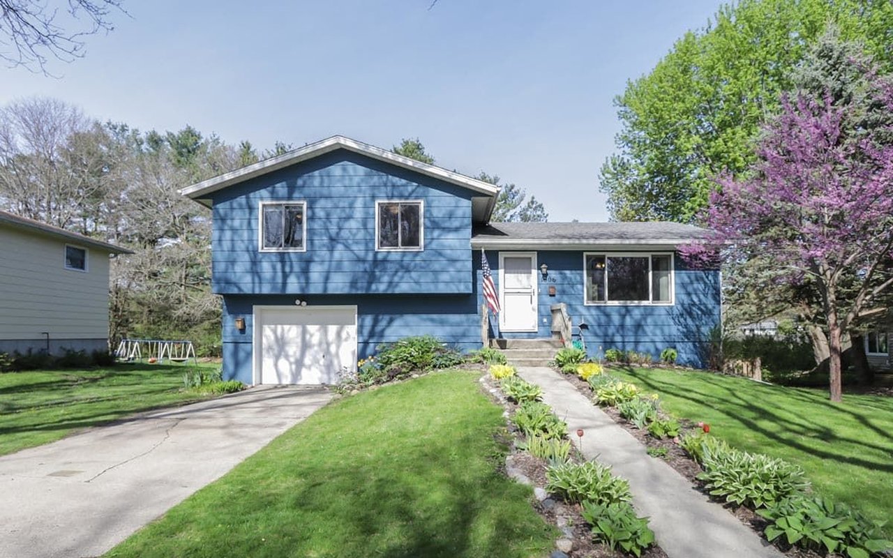 Blue split-level house with an attached garage, flower-lined walkway, and pink-blossoming tree.