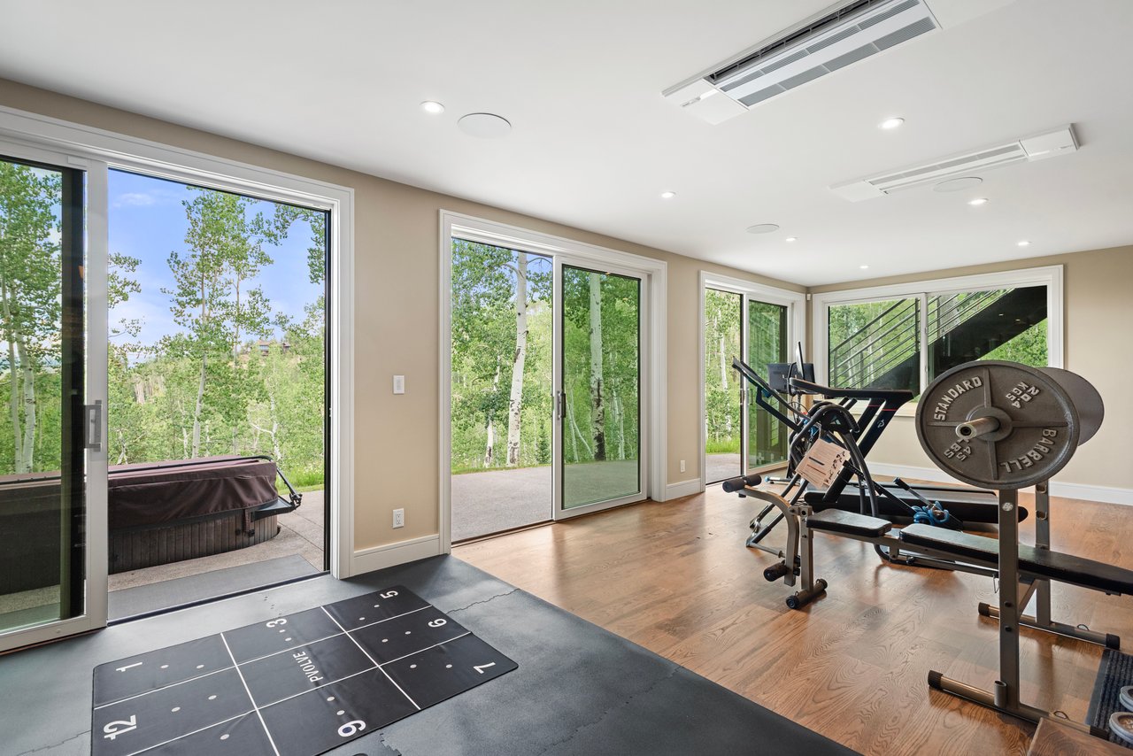 Home gym with weight equipment and a floor mat, opening onto a wooded patio with a hot tub.