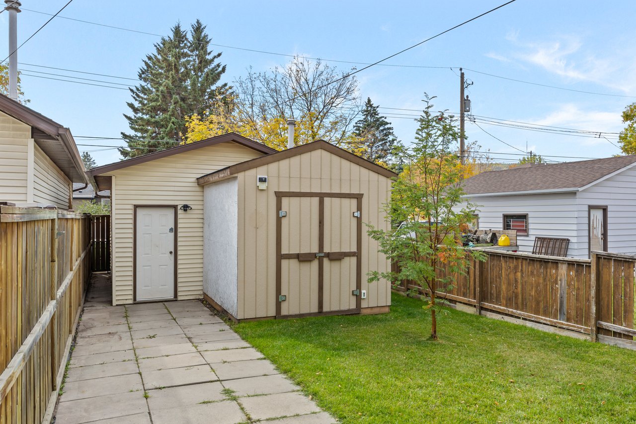Backyard with a tan storage shed beside a paved walkway and fenced lawn.