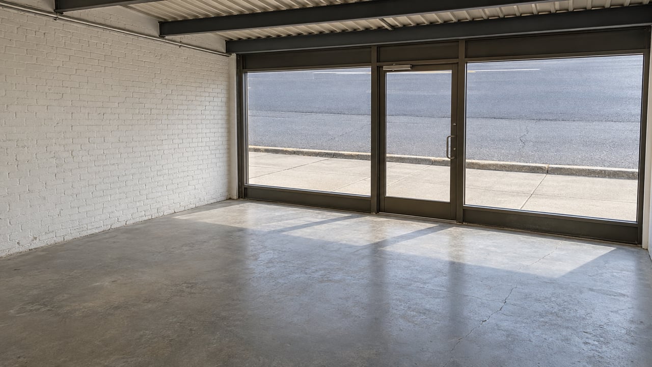 An unfurnished commercial interior with worn gray concrete flooring, painted brick, exposed steel ceiling structure and a closed glass storefront.