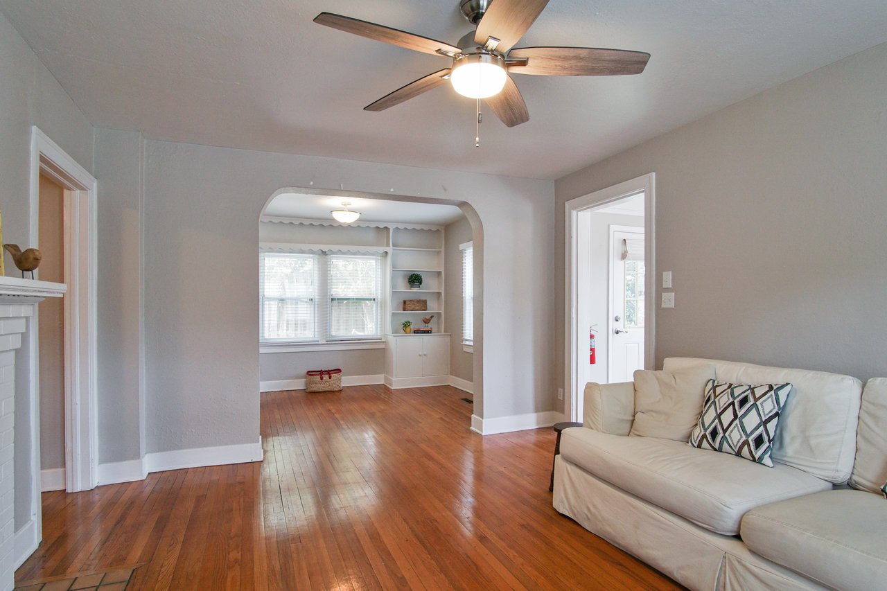 Living room with hardwood floors, white walls, and a ceiling fan. A white sofa with a patterned pillow is on the right, adjacent to an archway leading to a bright room with built-in shelves and large windows. Cozy atmosphere.
