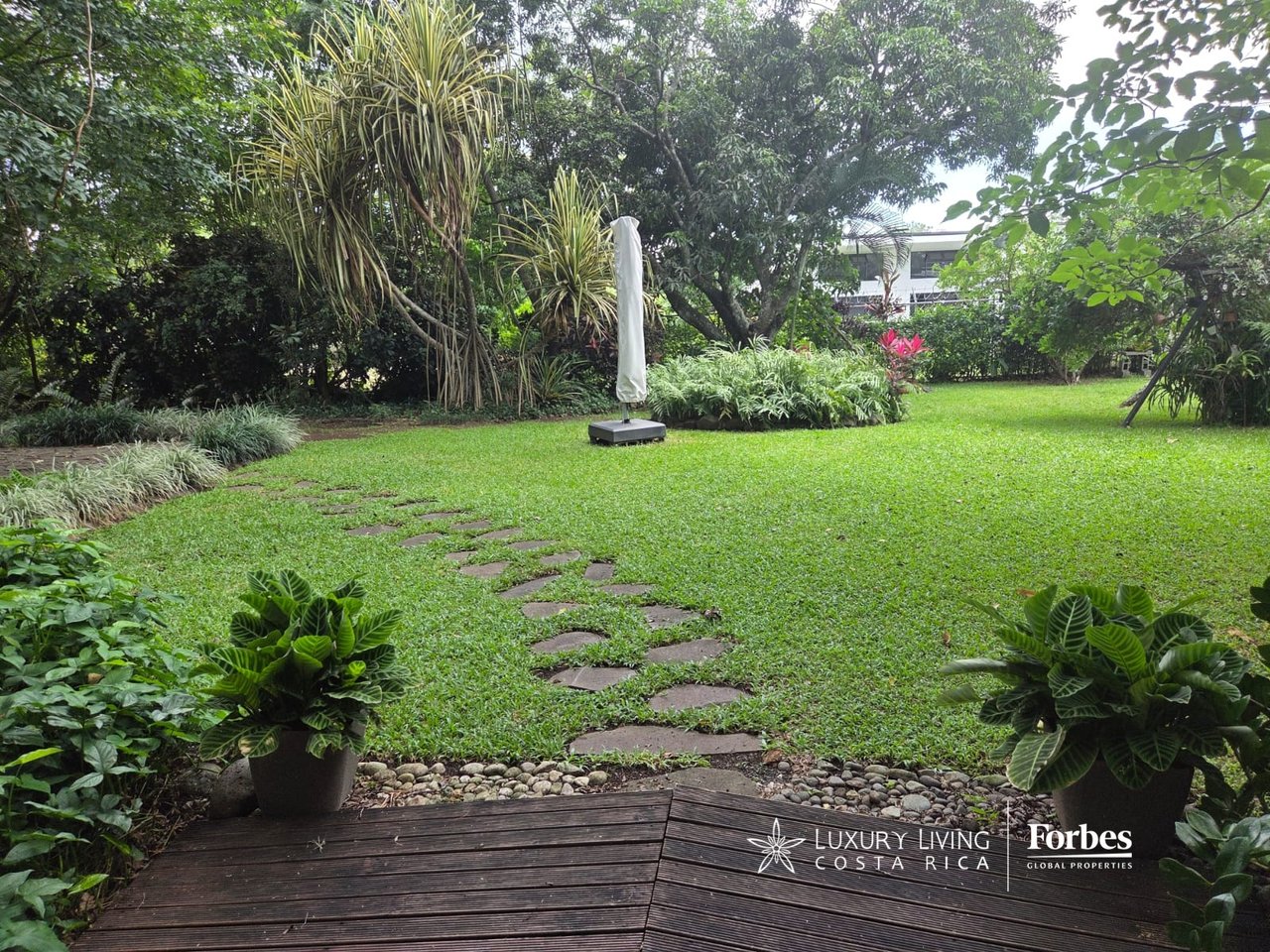 Villa garden with a broad lawn, stepping-stone path, tropical plants, and a wooden deck.