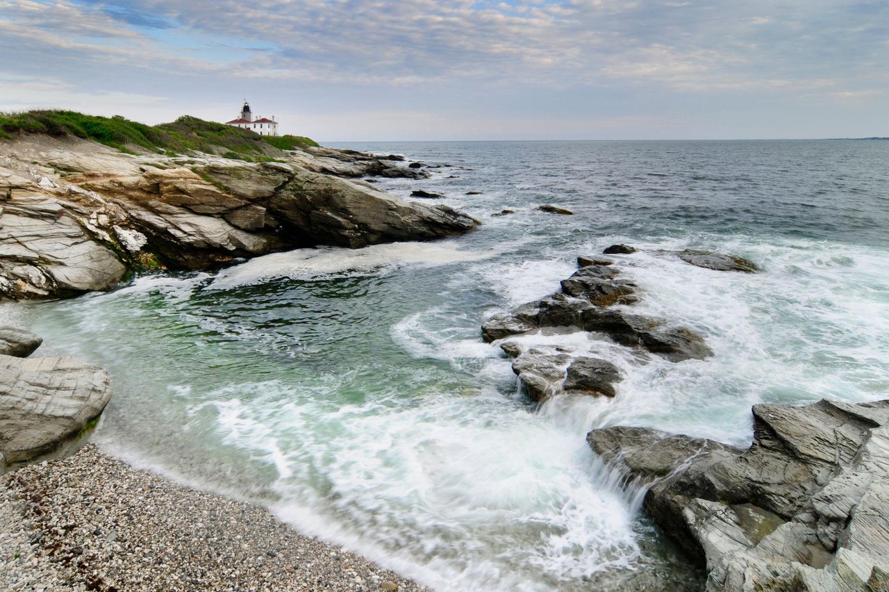 Rocky shoreline and foaming cove below a lighthouse on a grassy headland.