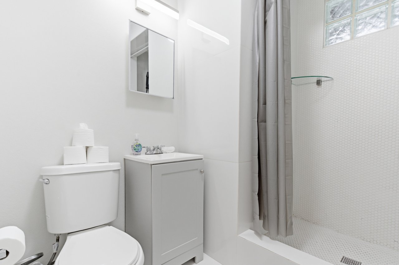 White bathroom with a compact vanity and gray-curtained shower beneath a glass-block window.