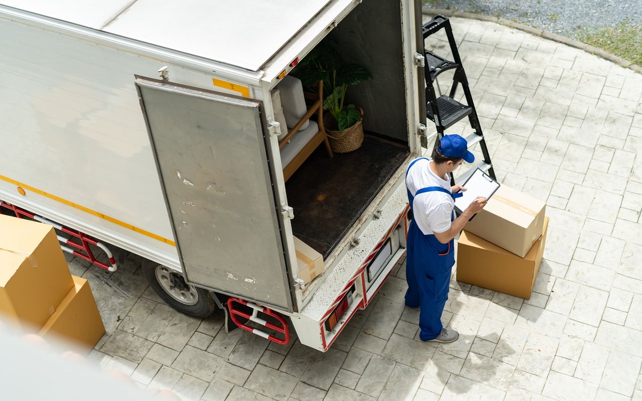 Mover checks a clipboard beside boxes and an open moving truck.