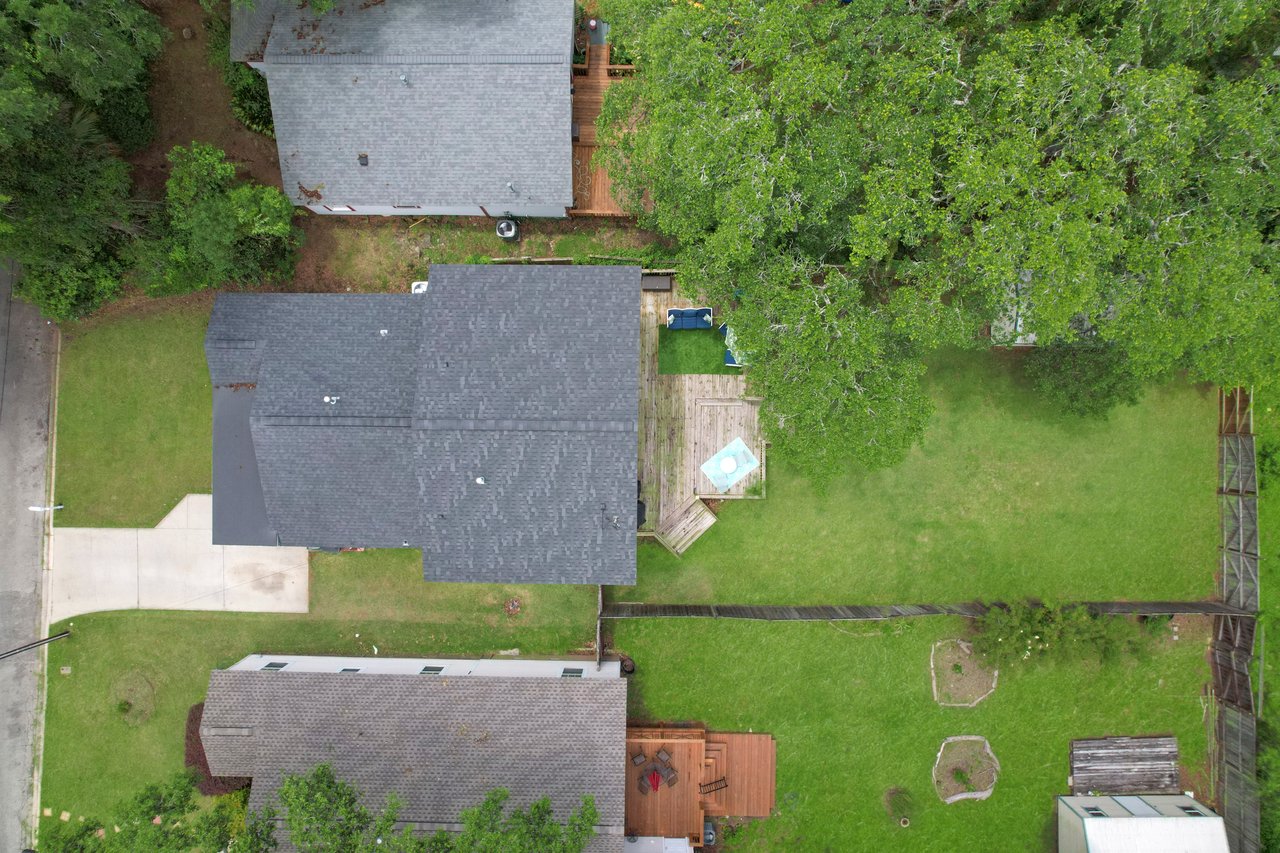 Aerial view of suburban backyard with green lawn, two gray-roofed houses, a small patio, and bordering trees. Calm, organized neighborhood scene.