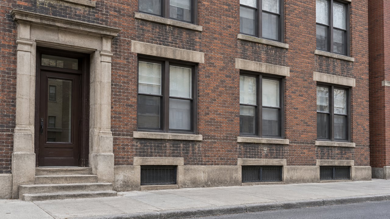 Weathered five-story brick walk-up with paired windows, pale stone lintels, and a recessed entrance above worn steps.