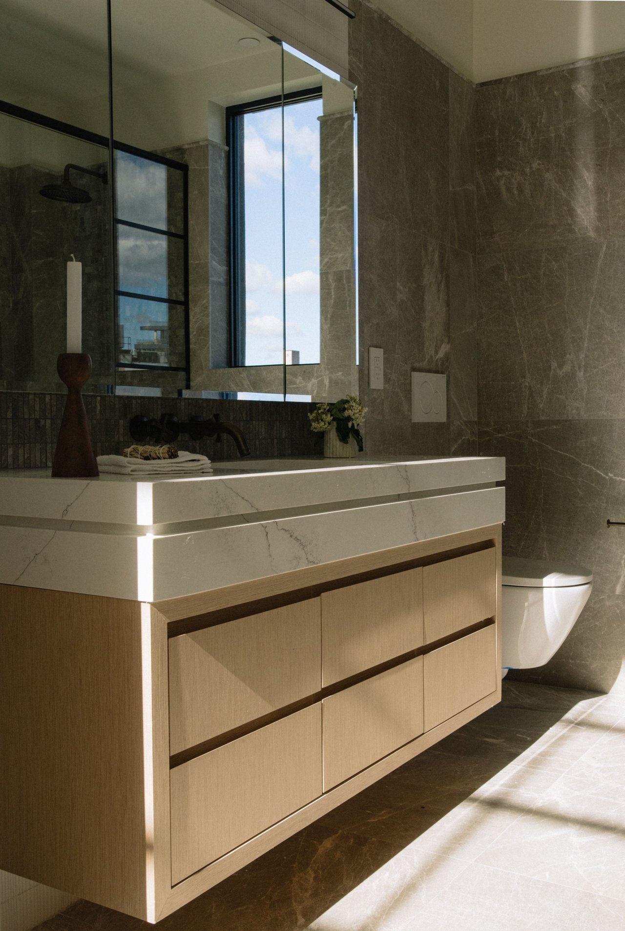 Modern bathroom with a light-wood vanity, veined stone walls, and a tall window.