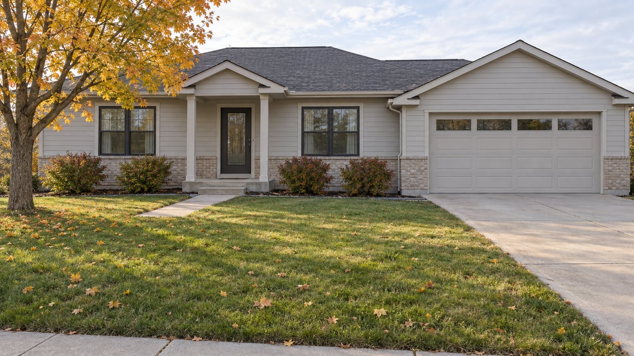 A light-gray single-story home with brick accents, a broad front lawn and a yellow-orange maple in soft autumn daylight.