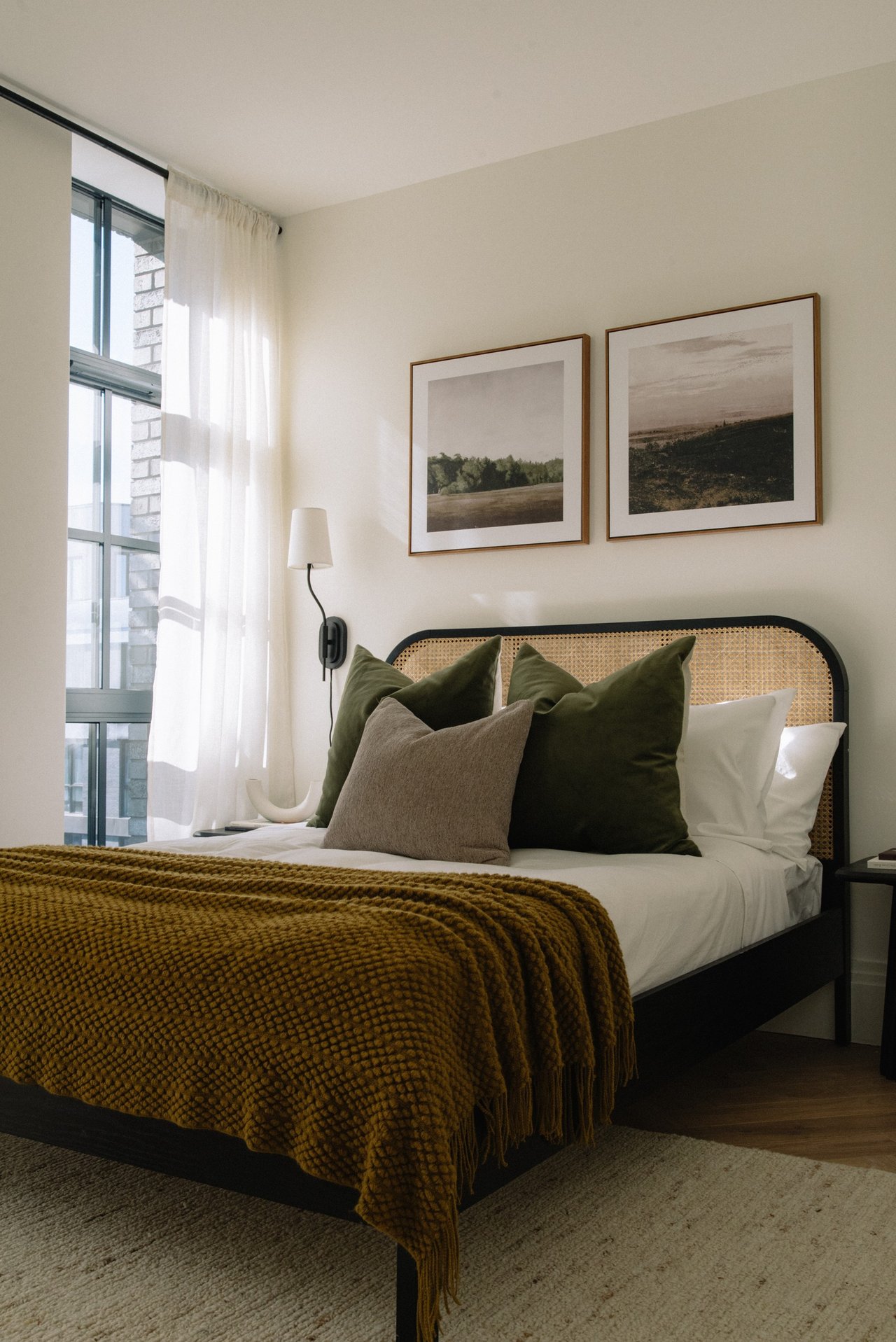 Bedroom with a cane headboard, olive pillows, and a mustard knit blanket beside tall curtained windows.