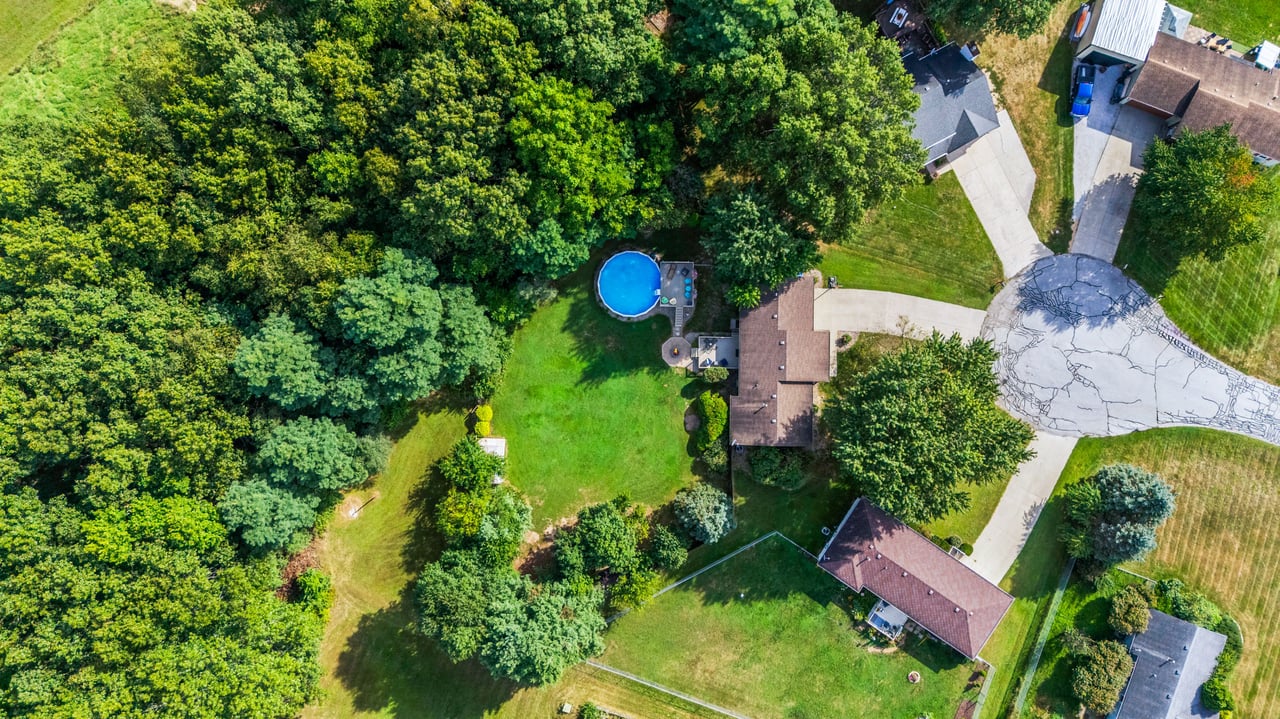 Aerial view of a wooded property with a broad lawn, blue above-ground pool, and curved driveway.