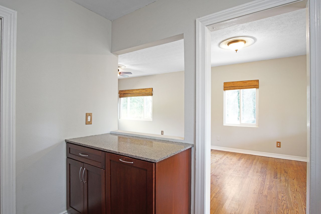 Hallway view featuring a dark wood cabinet with a granite countertop, flanked by two rooms with white walls, wood floors, and bright windows.