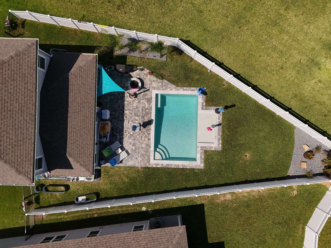 Aerial view of a backyard pool and paved patio surrounded by lawn and a white fence.