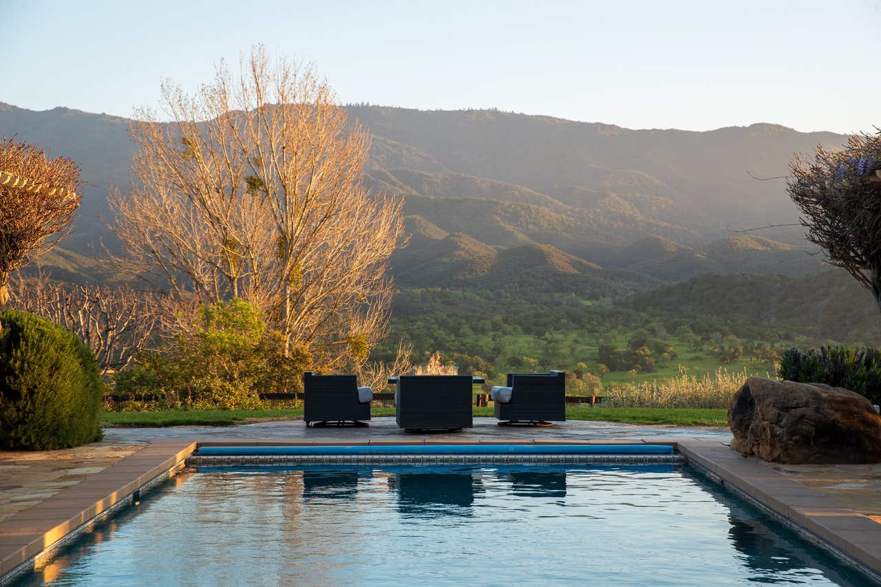 Swimming pool overlooking a green valley and mountains, with three lounge chairs on the stone patio.