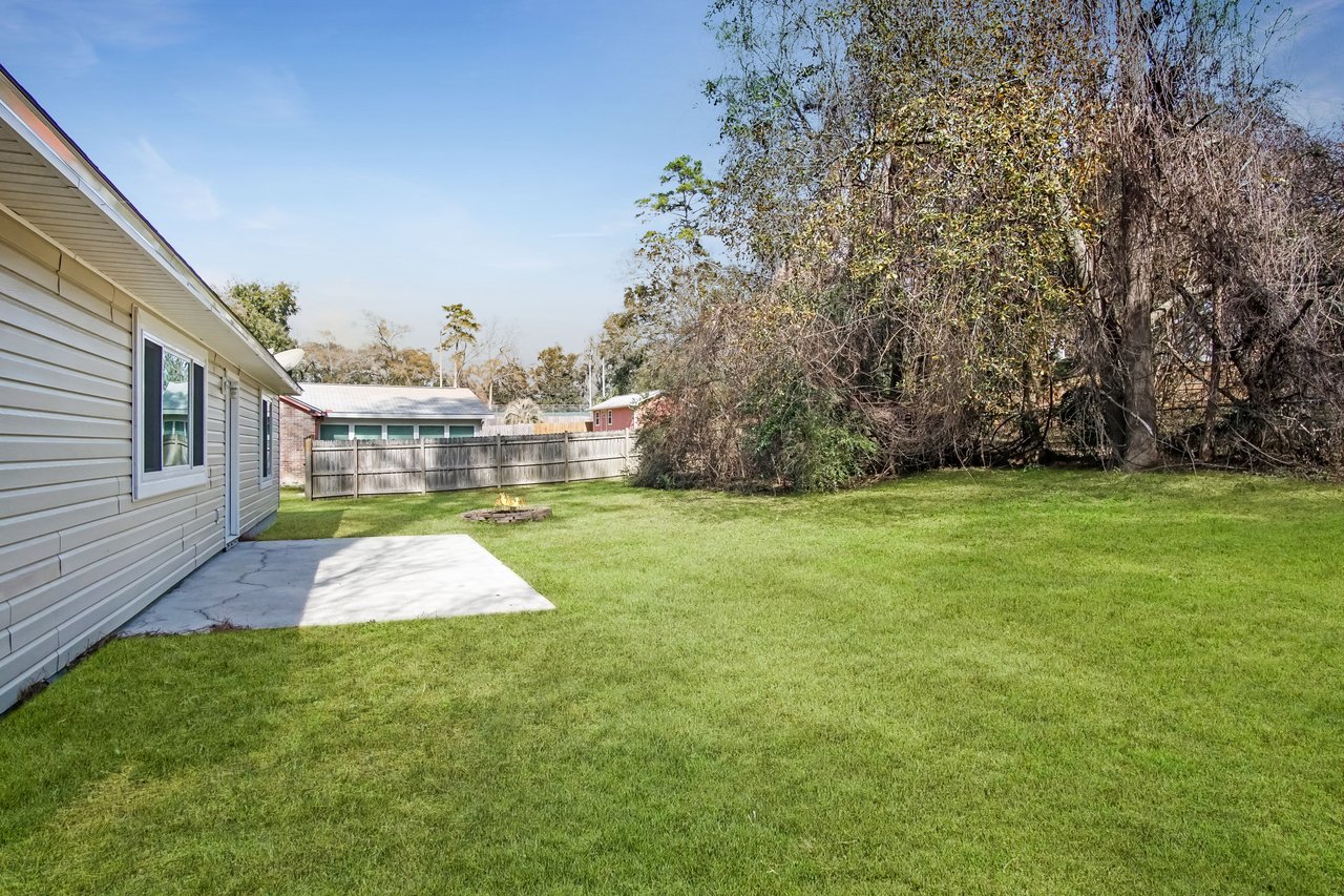 A backyard with green grass, a circular stone fire pit with a small flame, a beige house with large windows, and bare trees under a clear sky.