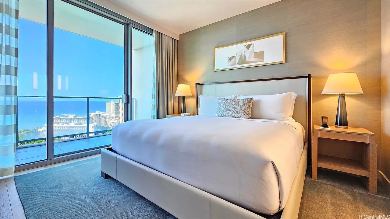 Bedroom with a white bed and balcony doors overlooking the ocean and city.