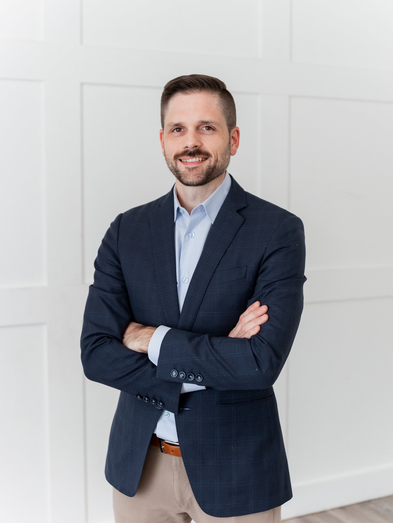 Paul Myers in a navy suit, standing with his arms crossed against a white paneled wall.