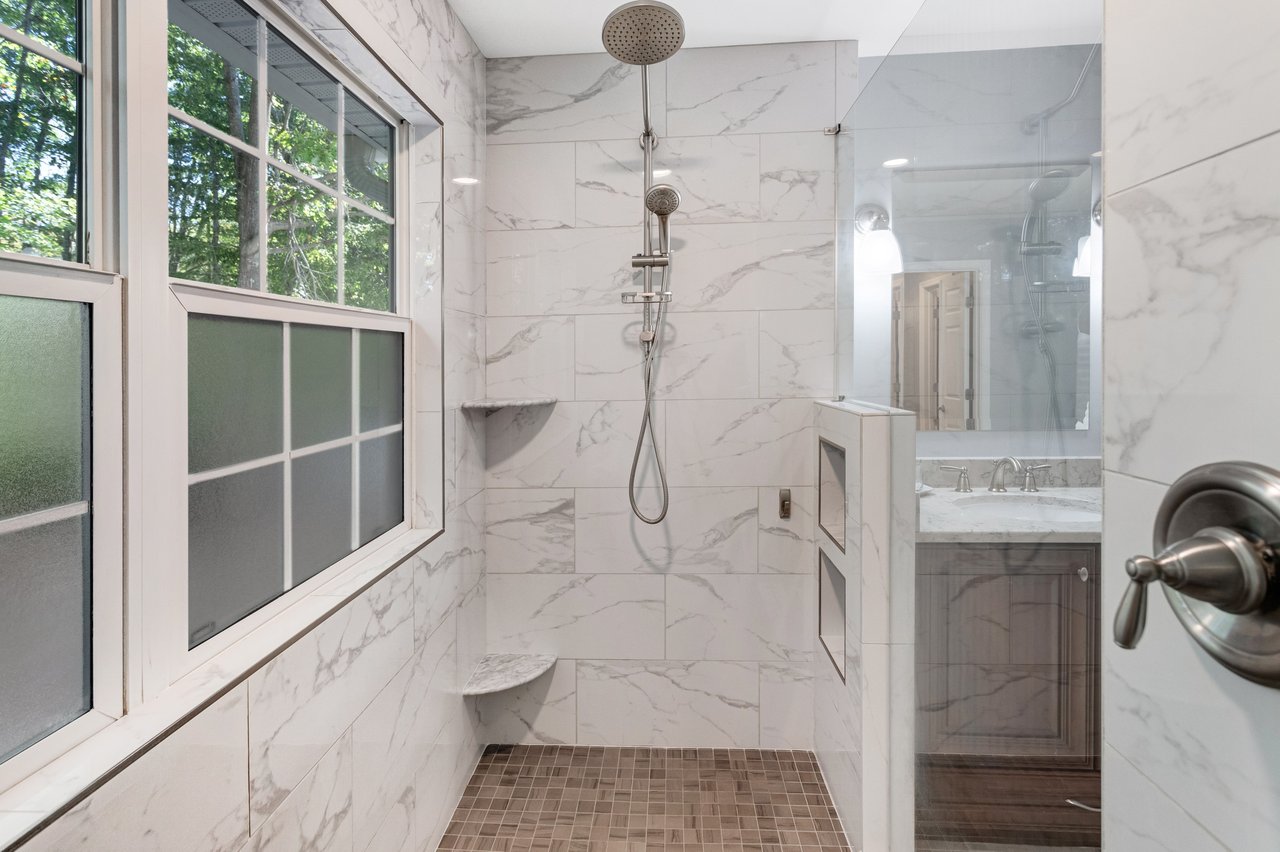 Walk-in marble-look tiled shower with a rainfall showerhead, glass partition, and large window.