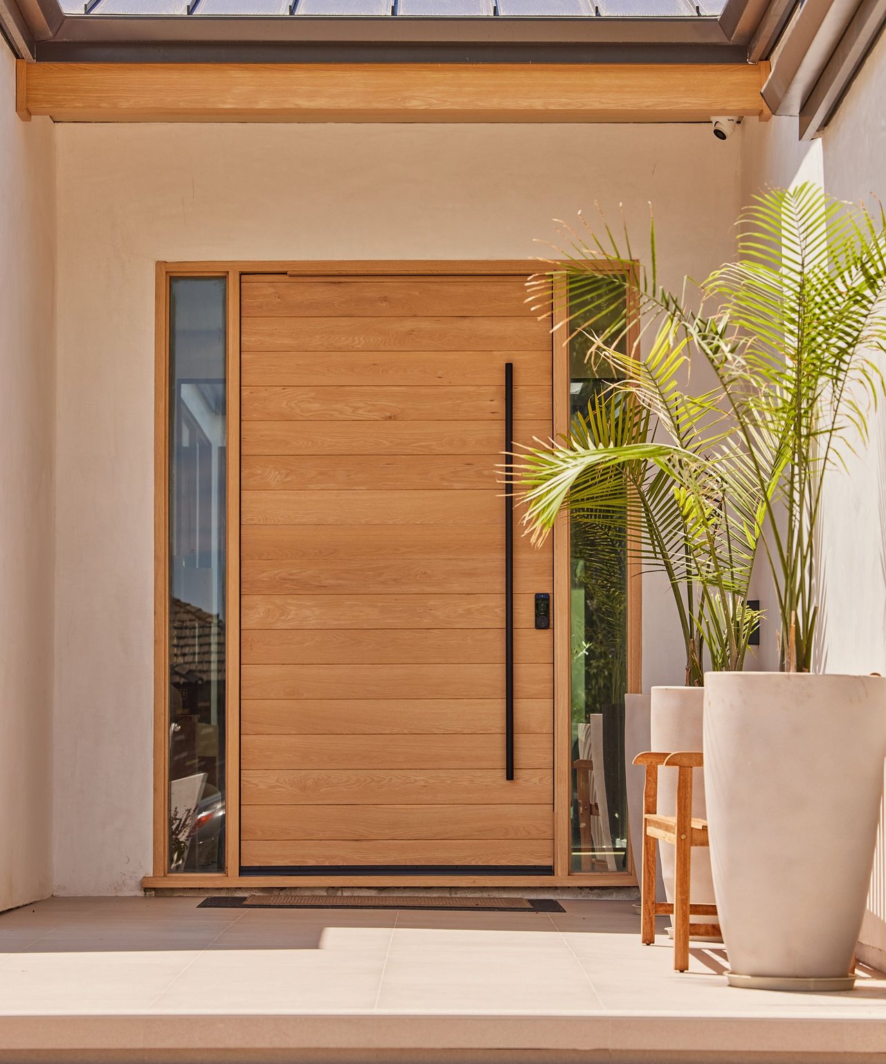 Modern wooden front door with glass sidelights, a skylight, and potted palms on the porch.