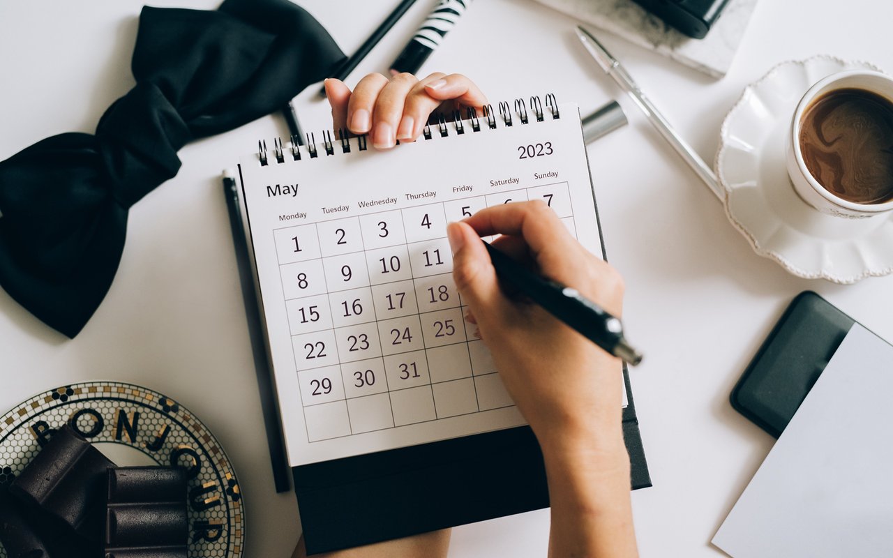A hand writes on a May 2023 desk calendar beside a cup of coffee.