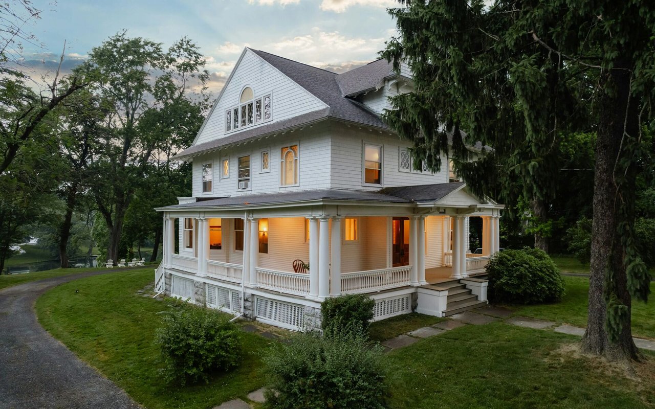 White Victorian-style house with a wraparound porch, glowing windows, and a wooded lawn.