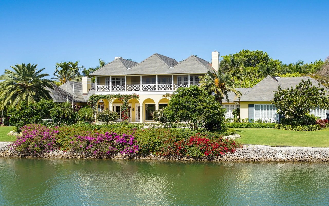 Two-story waterfront home with an arched veranda, palm trees, and colorful gardens beside the water.