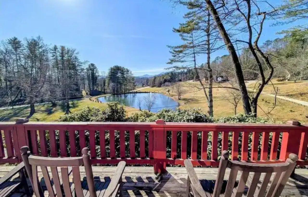 Deck overlooks a pond and open grounds, framed by a red railing and wooden chairs.