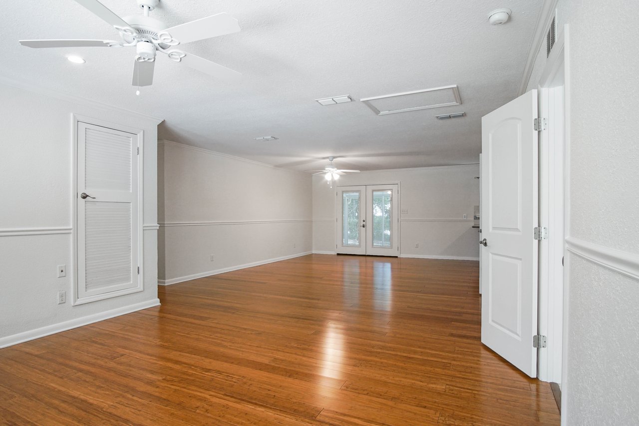 Spacious empty room with wooden flooring, white walls, and a ceiling fan. Double doors with glass panels lead outside, creating a bright, open atmosphere.
