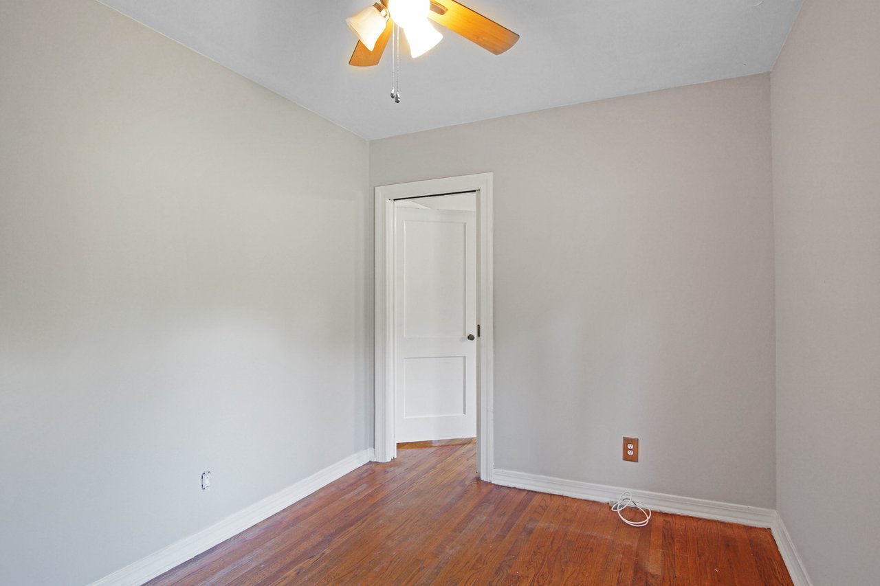 Empty room with light gray walls, a white door, and a ceiling fan with wooden blades. The room has a warm wooden floor and a relaxed, neutral vibe.
