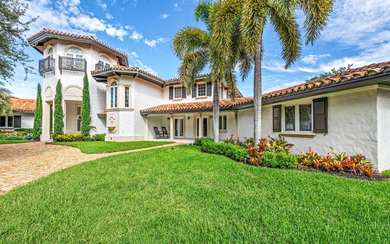White stucco home with terracotta roofs, wrought-iron balconies, and palms beside a broad green lawn.