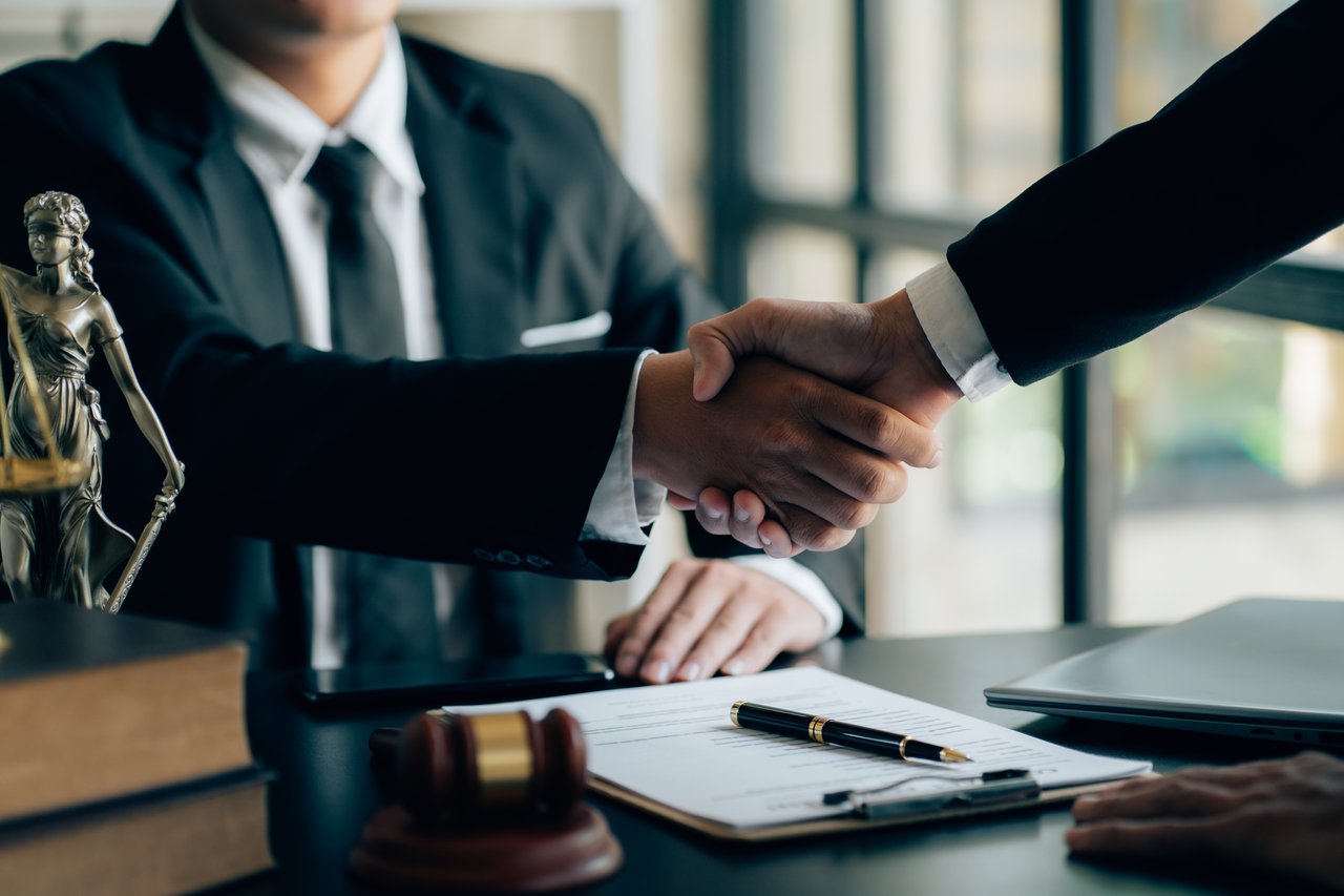 Two individuals in suits shake hands across a desk after closing a real estate deal