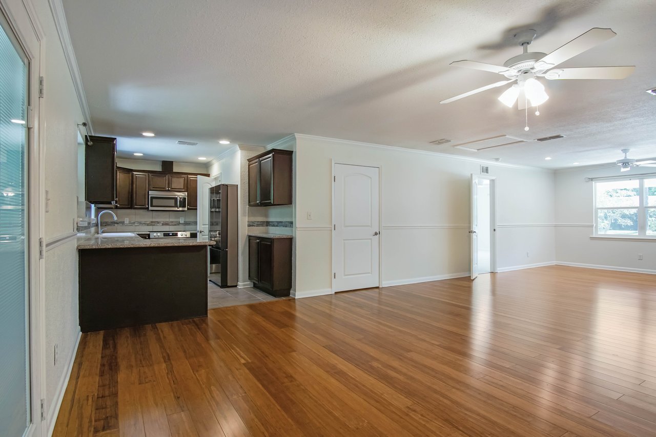 Spacious living area with hardwood floors and ceiling fan. Open kitchen features dark cabinets and stainless steel appliances. Bright natural light.