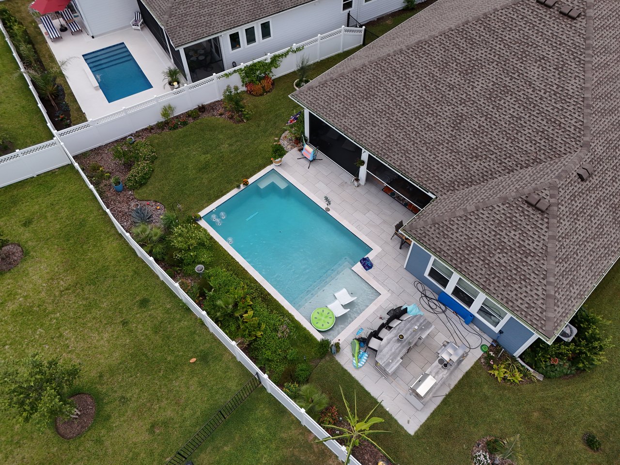 Aerial view of a fenced home backyard with a swimming pool, stone patio, outdoor seating, and garden beds.