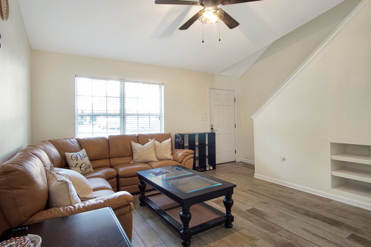Cozy living room with a brown leather sectional, glass coffee table, and wall shelves. Bright window and light fan create an airy atmosphere.Cozy living room with a brown leather sectional, glass coffee table, and wall shelves. Bright window and light fan create an airy atmosphere.