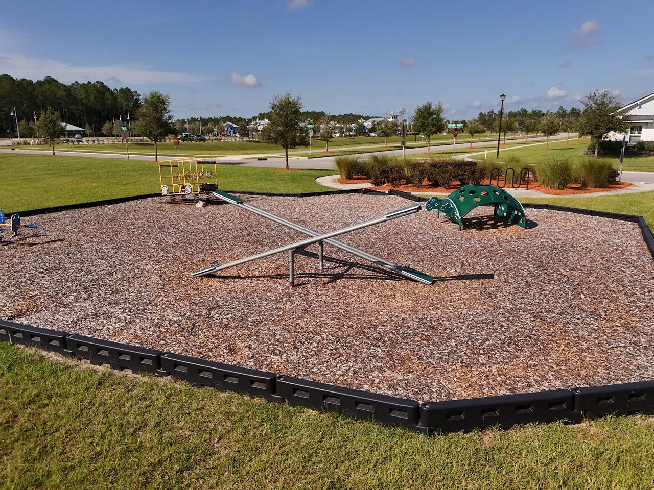 Community playground with a wood-chip surface, seesaw, and green climbing equipment.