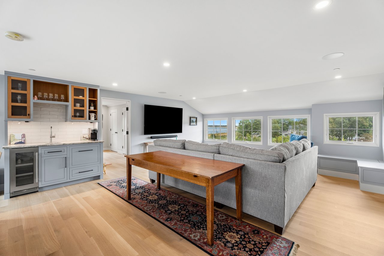 Living room with gray sectional, built-in kitchenette, and windows overlooking the water.