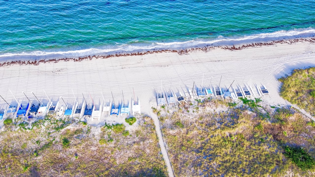 Aerial beach with turquoise waves, white sand, and rows of small sailboats.
