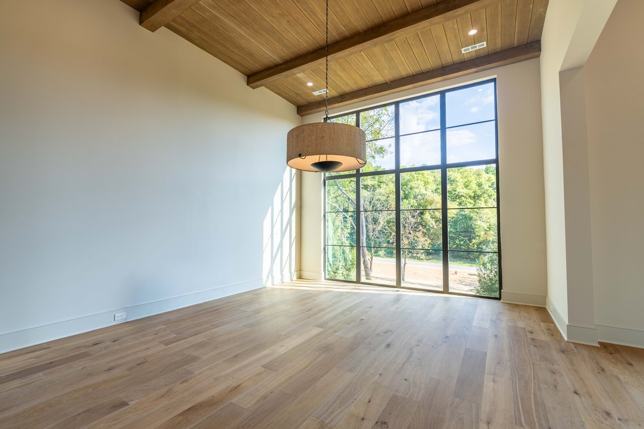 Empty room with exposed ceiling beams, hardwood floors, and large windows overlooking trees.