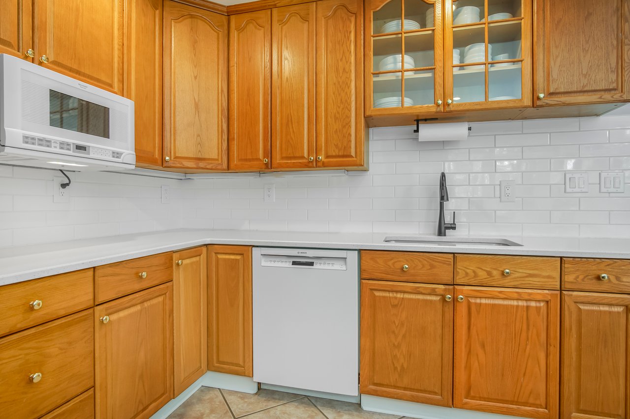 A cozy kitchen with wooden cabinets and white countertops. Features a microwave, dishwasher, and a tiled backsplash, creating a warm and inviting space.