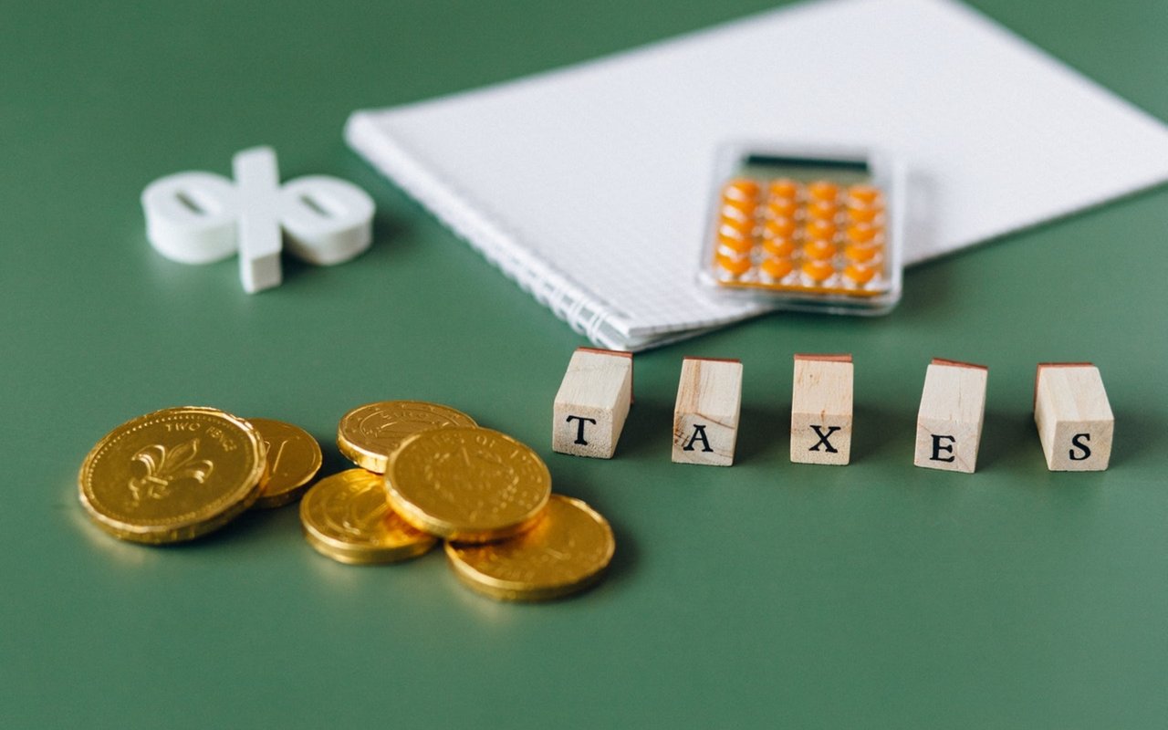 Wooden blocks spelling TAXES beside gold-colored coins, a percent symbol, notebook, and calculator.